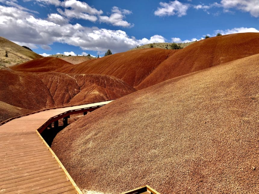 A wooden boardwalk winds through the colorful, rolling hills of the Painted Hills, Oregon, under a sky with scattered clouds.