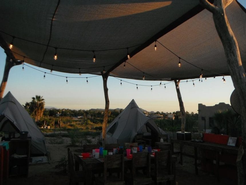 Outdoor area with tents, a shaded canopy, string lights, and rustic wooden furniture. Scenic view with palm trees in the background.