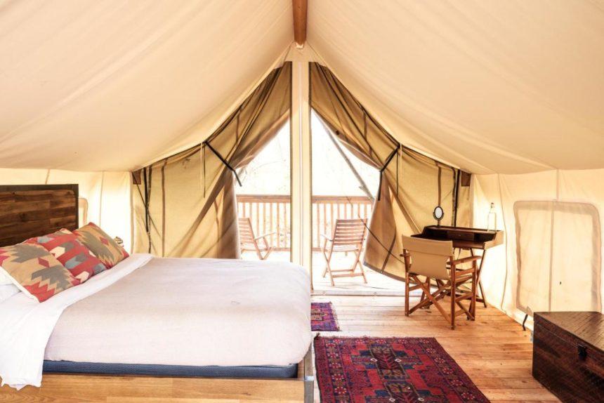 A cozy glamping tent interior features a bed, wooden floor, seating area, and rugs. The tent opens to a wooden deck with two chairs.